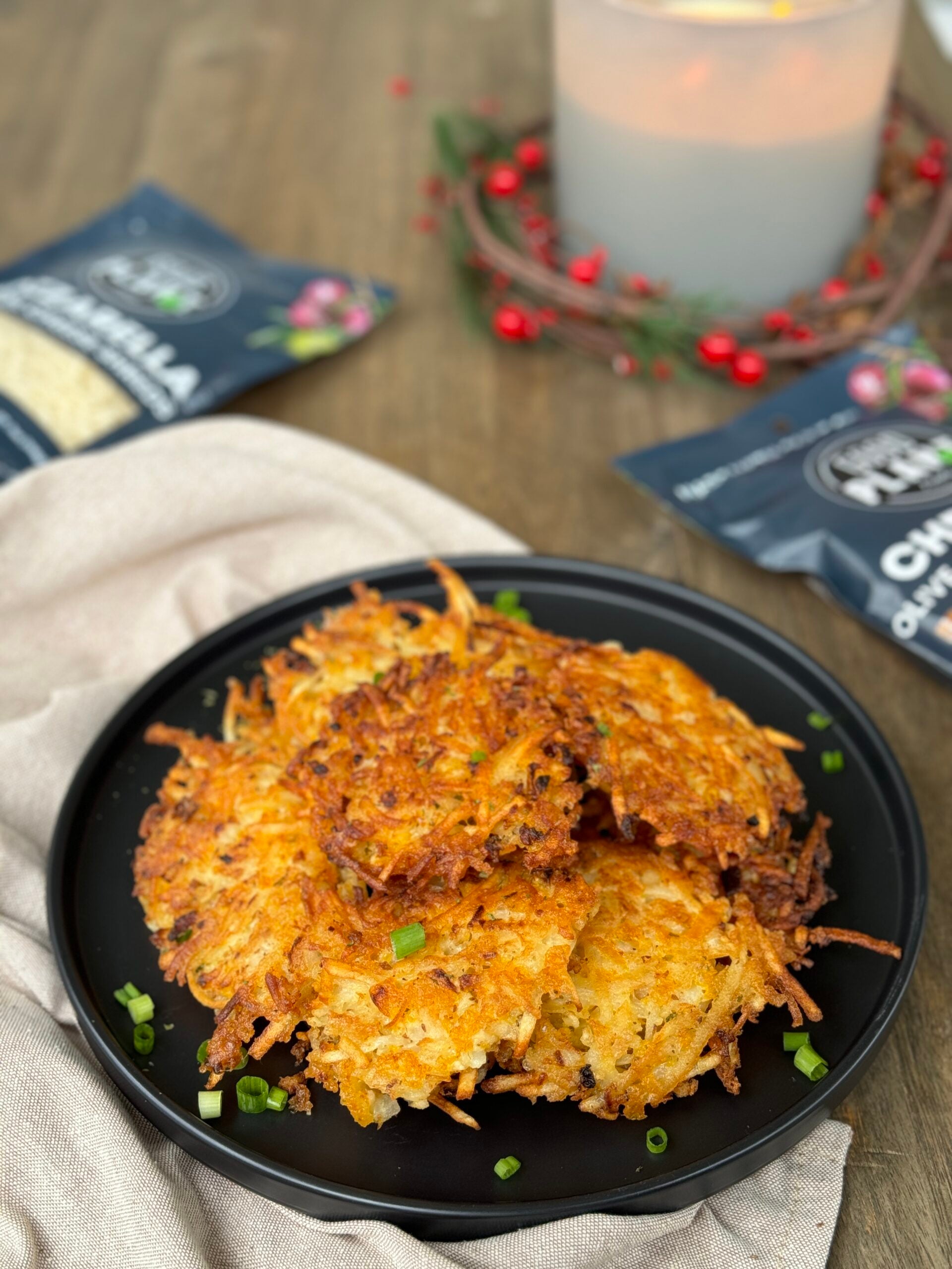 A plate of golden, crispy potato and cheese latkes stacked on top of each other, with melted cheese oozing out from the edges. The latkes are perfectly fried to a crunchy texture, made with olive oil and cheese. In the background, there are several bags of shredded cheese placed nearby, adding to the cheesy theme of the dish.